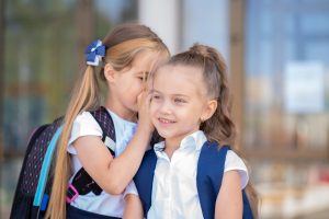 Two schoolgirls standing outside a school building with their heads physically close as one whispers a secret into the other's ear