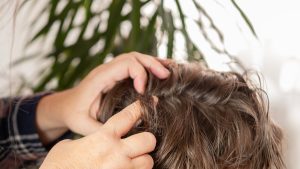 Parent's hands carefully parting a school-aged child's hair near the scalp to check for lice or dandruff under bright bathroom light