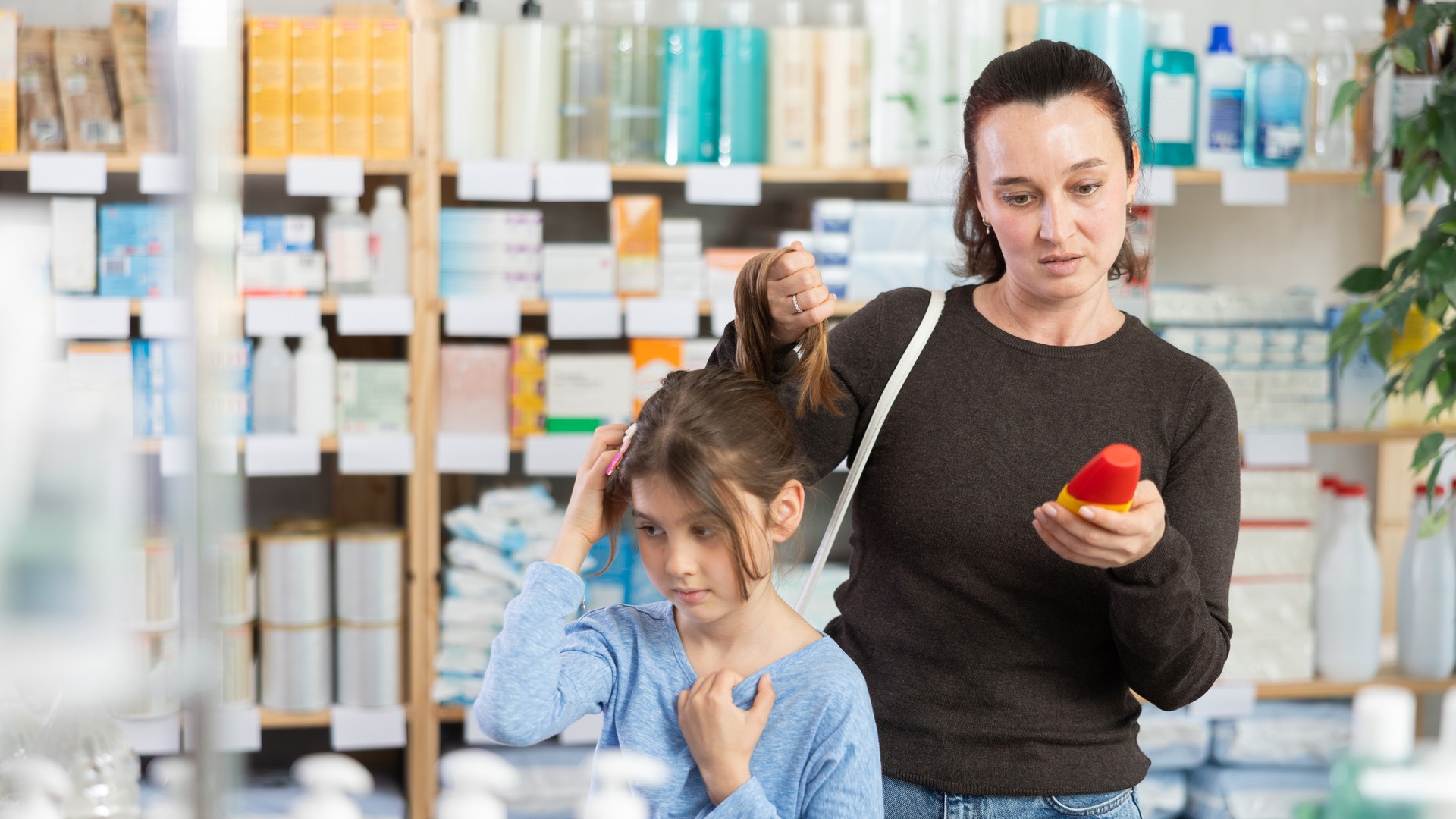 A concerned parent parting her daughter's hair and reading the label on a lice-remedy product while deciding how to treat a suspected case of head lice.