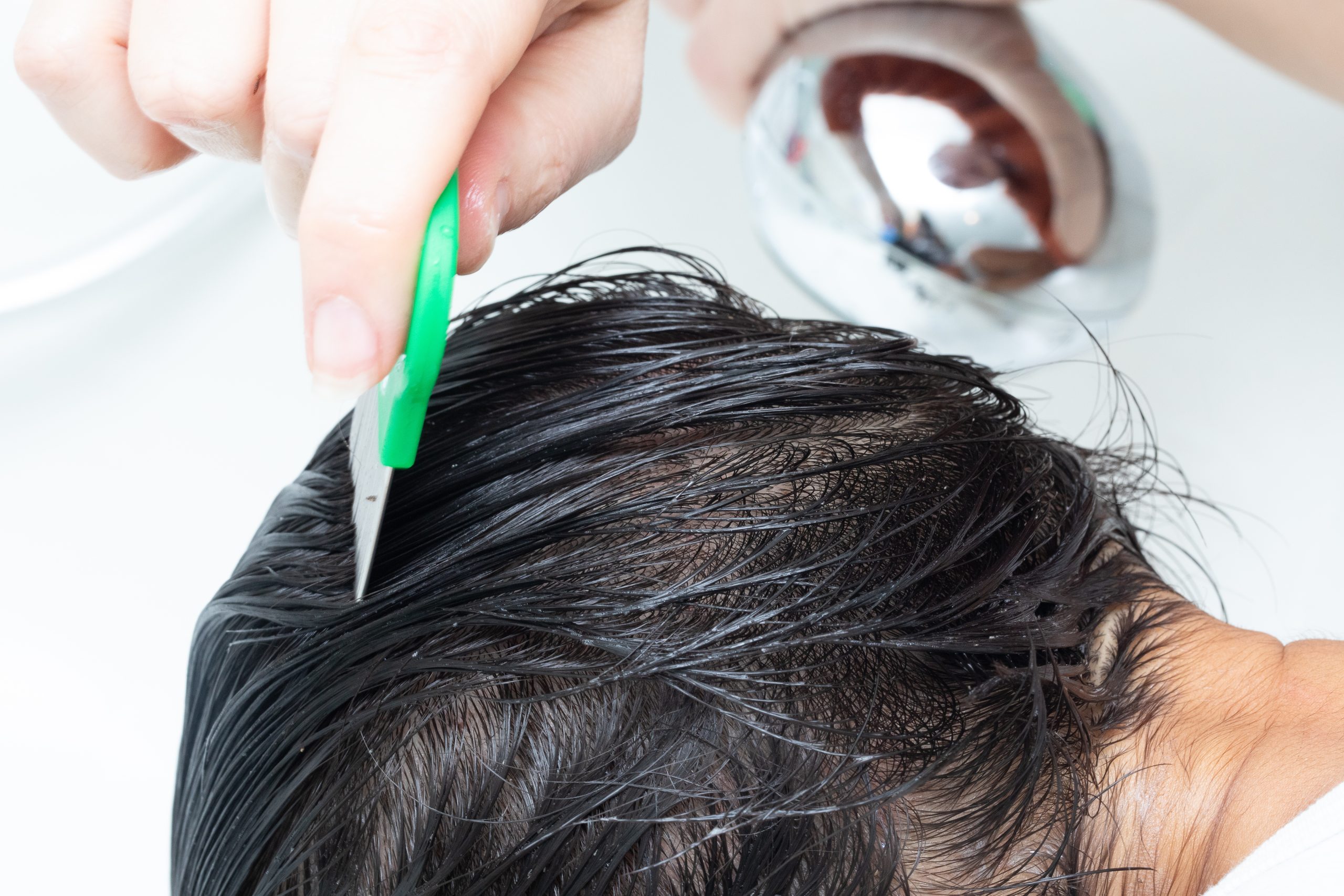 A parent sectioning a child's dry hair with a fine-tooth comb while applying a clear lice treatment lotion at a bathroom counter.