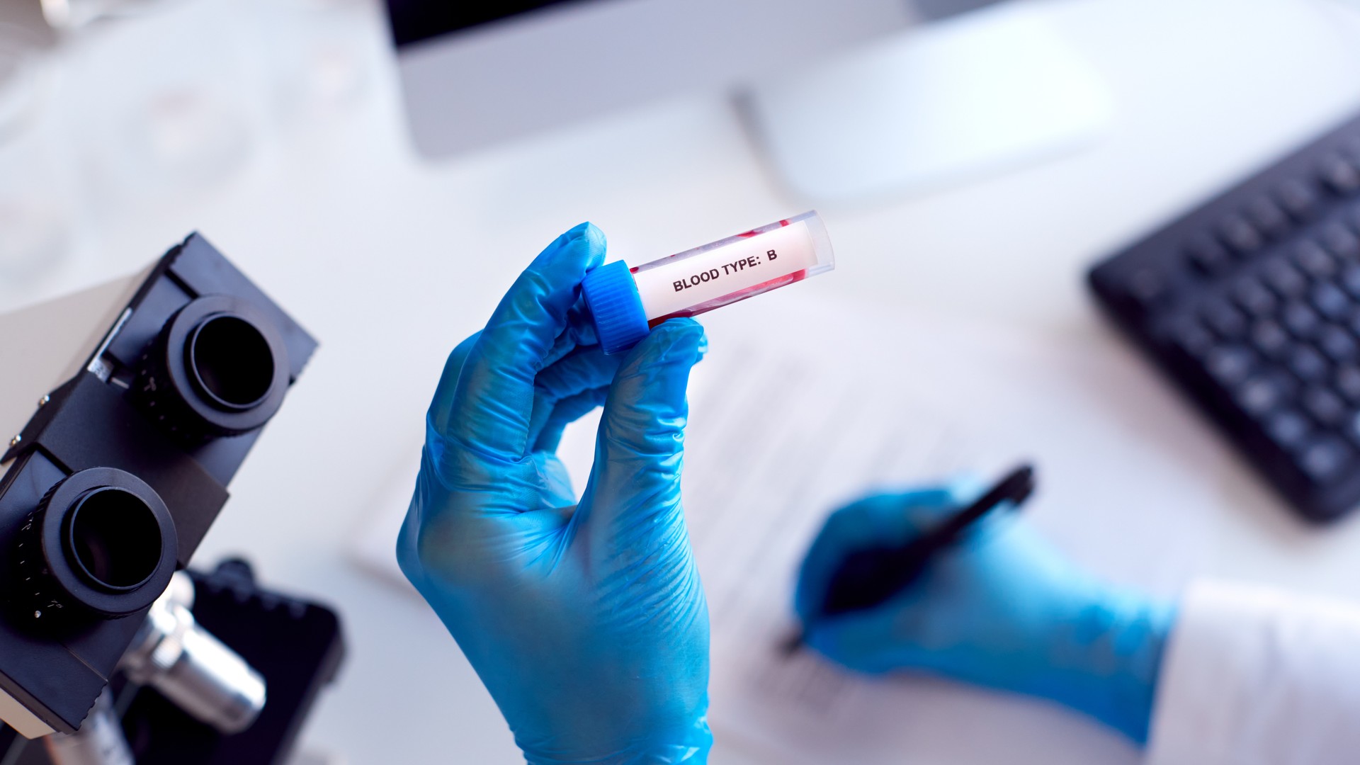 Laboratory worker holding a labeled blood type sample beside a microscope