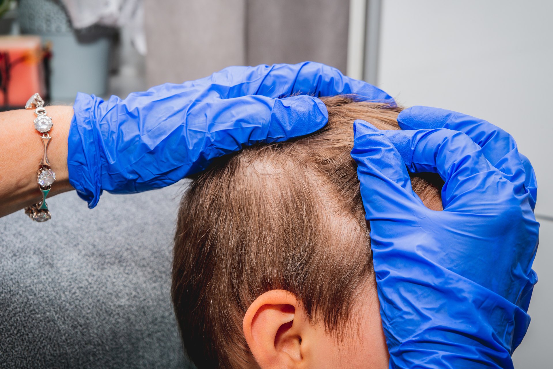 Gloved hands parting a fair-haired child's hair behind the ear to check the scalp