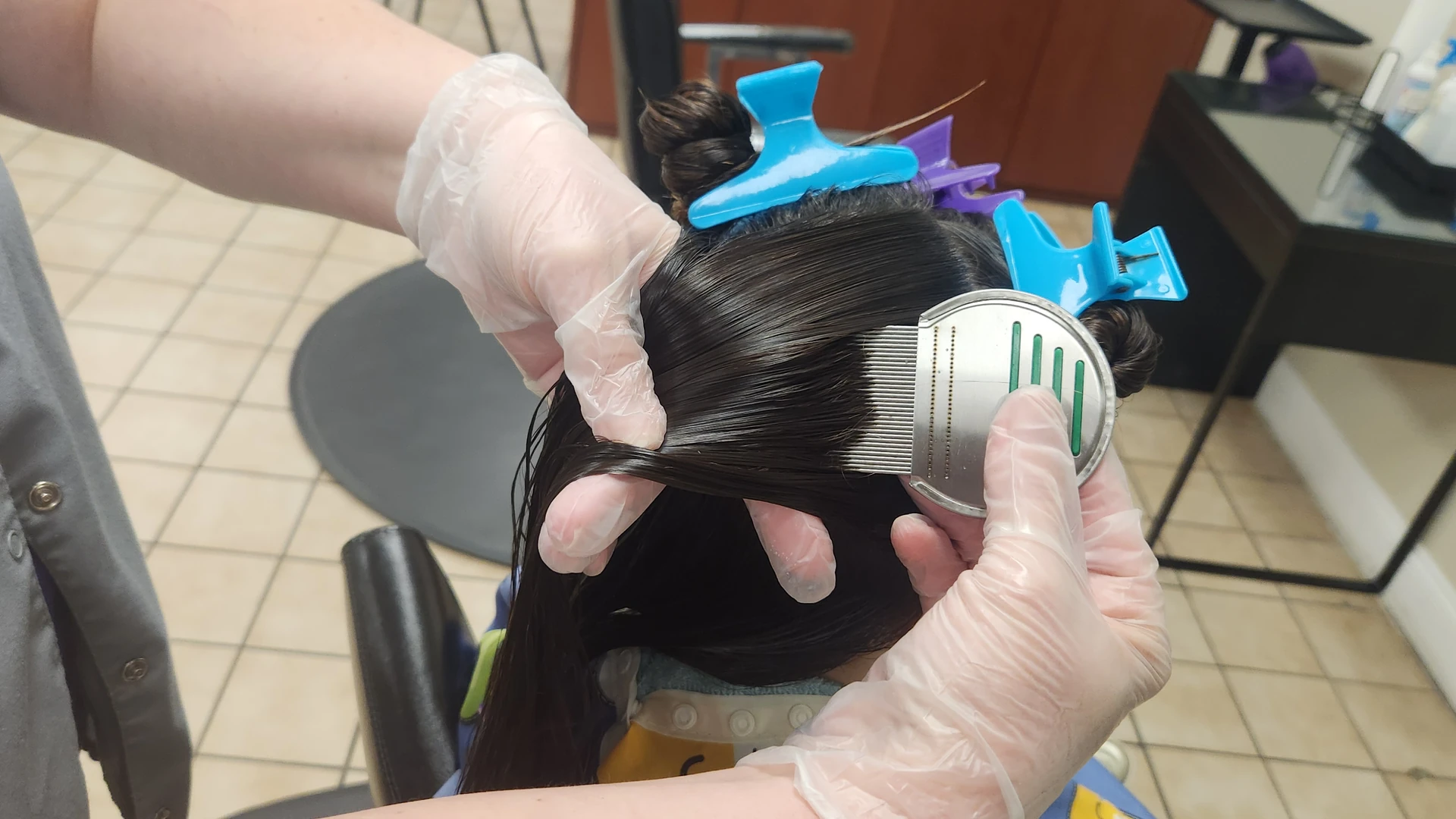 Gloved hands passing a fine-tooth comb through dark hair divided with blue and purple clips.