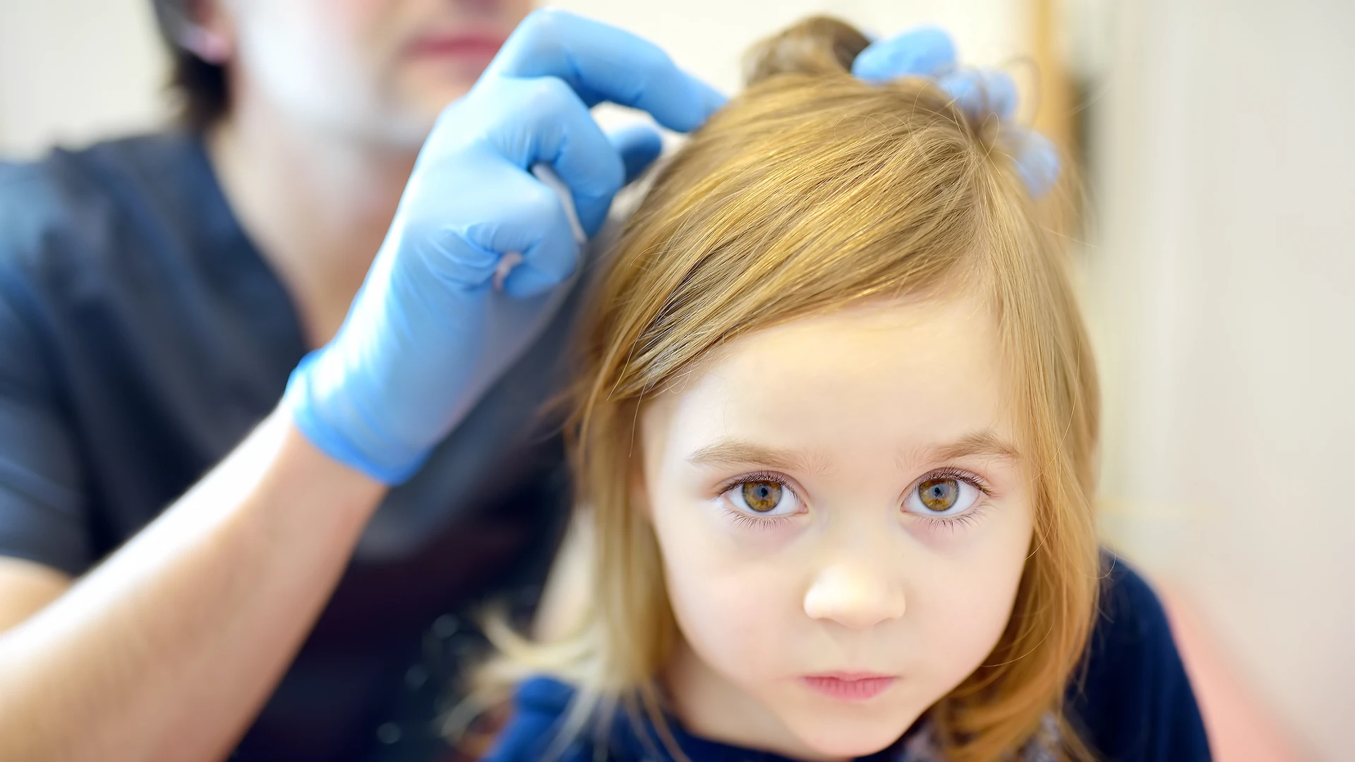 Person facing forward as blue-gloved hands part their hair near the crown.